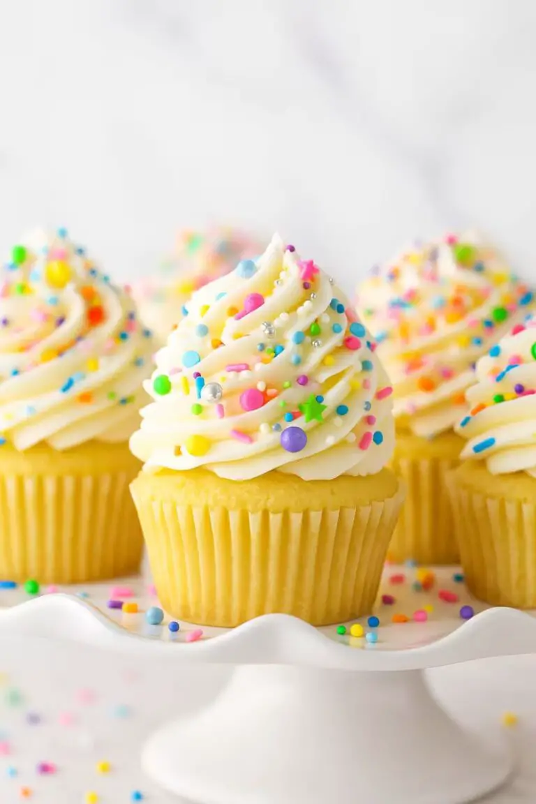 Vanilla cupcakes with creamy white frosting topped with colorful rainbow sprinkles on a cake stand.