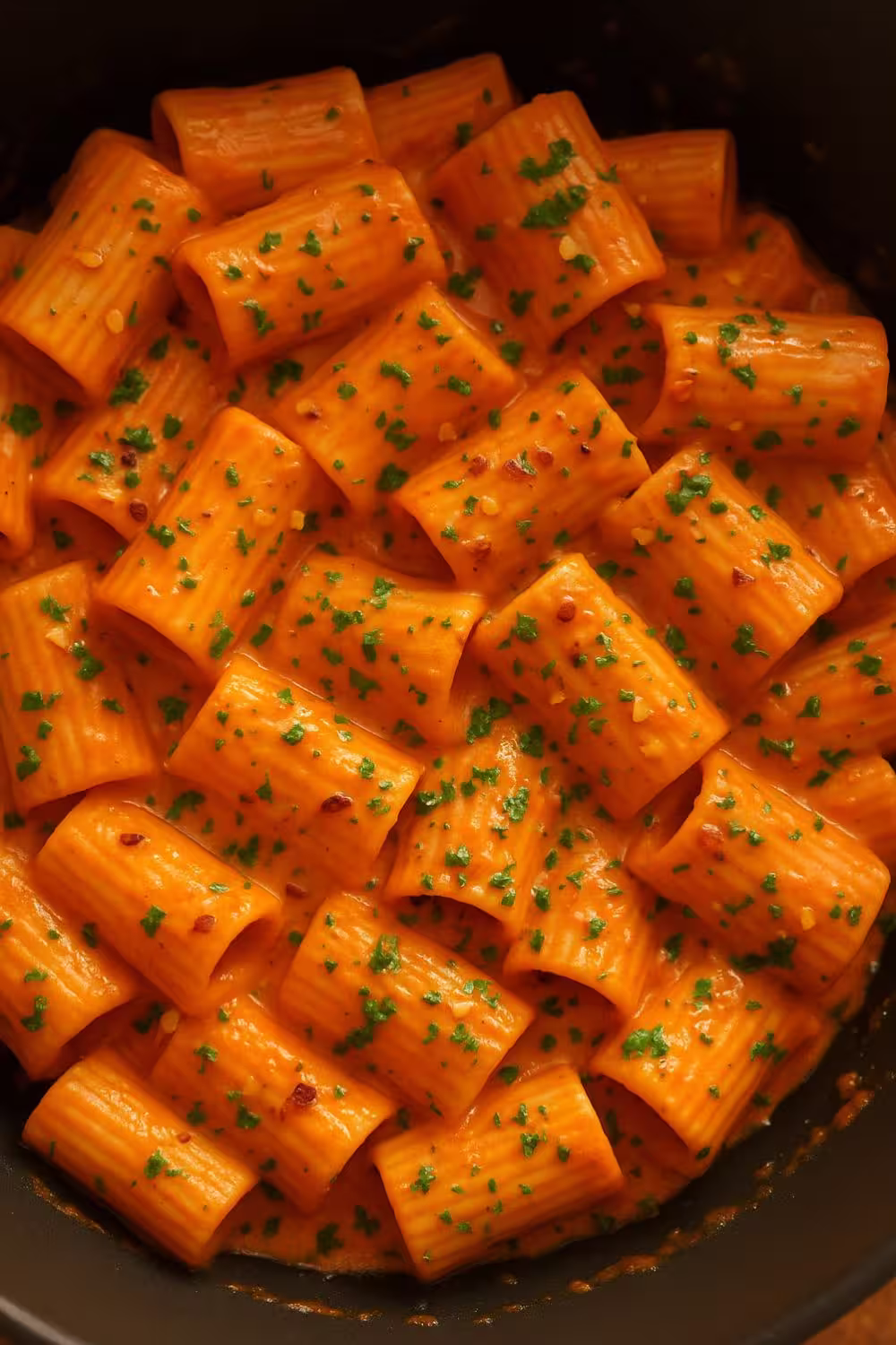 Creamy spicy rigatoni pasta topped with red pepper flakes and chopped parsley in a skillet.