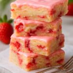 Stack of moist strawberry lemon blondies topped with pink glaze, sitting next to forks.