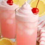 A tall glass of whipped pink lemonade topped with a swirl of creamy foam, garnished with a maraschino cherry and a slice of lemon.