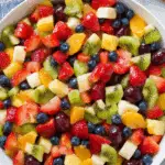 Honey Lime Rainbow Fruit Salad with strawberries, kiwi, bananas, blueberries, grapes, oranges, and pineapple in a white bowl.