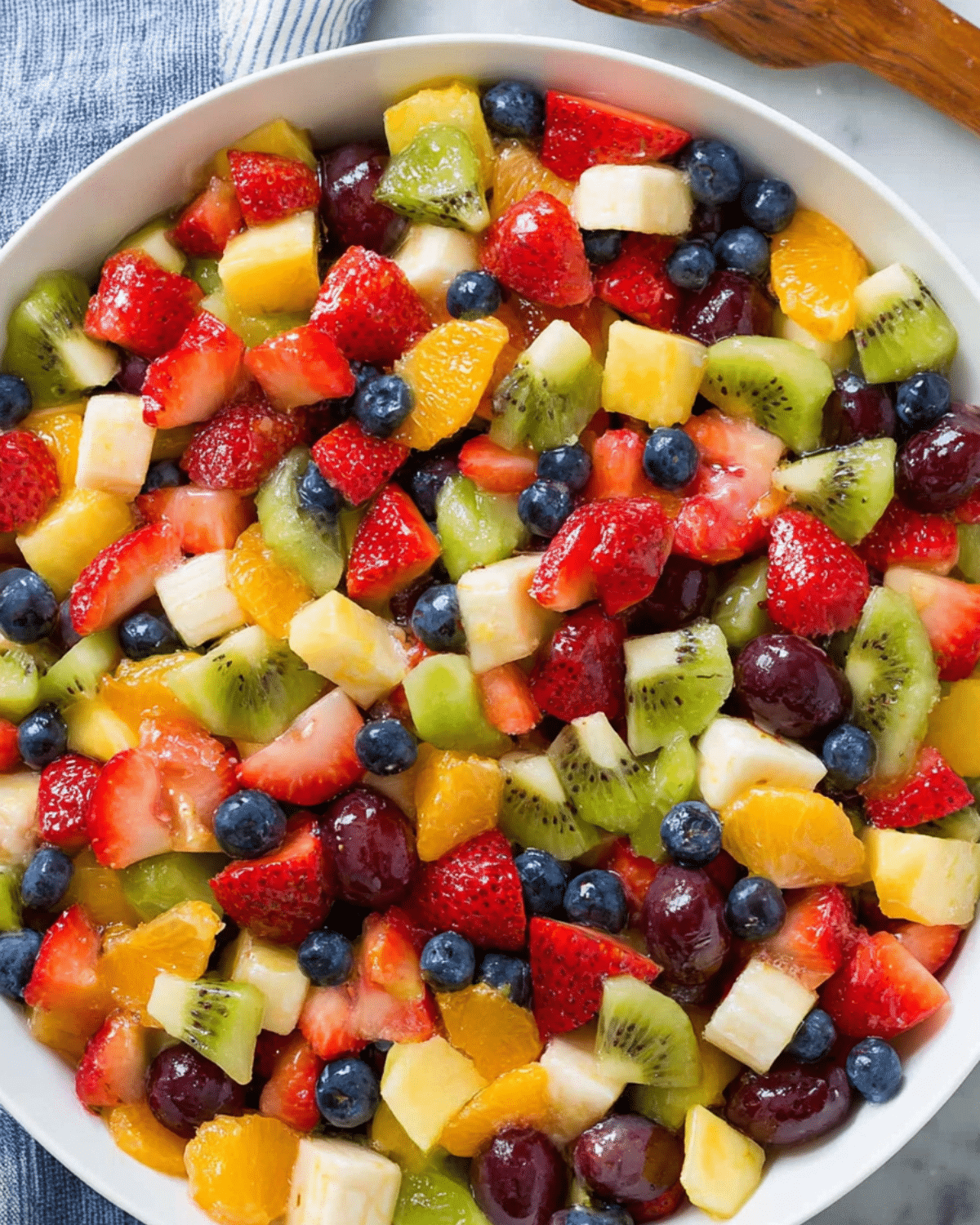 Honey Lime Rainbow Fruit Salad with strawberries, kiwi, bananas, blueberries, grapes, oranges, and pineapple in a white bowl.