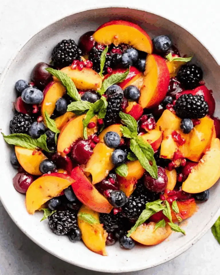 A bowl of summer peach fruit salad with fresh peach slices, blueberries, blackberries, cherries, pomegranate seeds, and mint leaves.