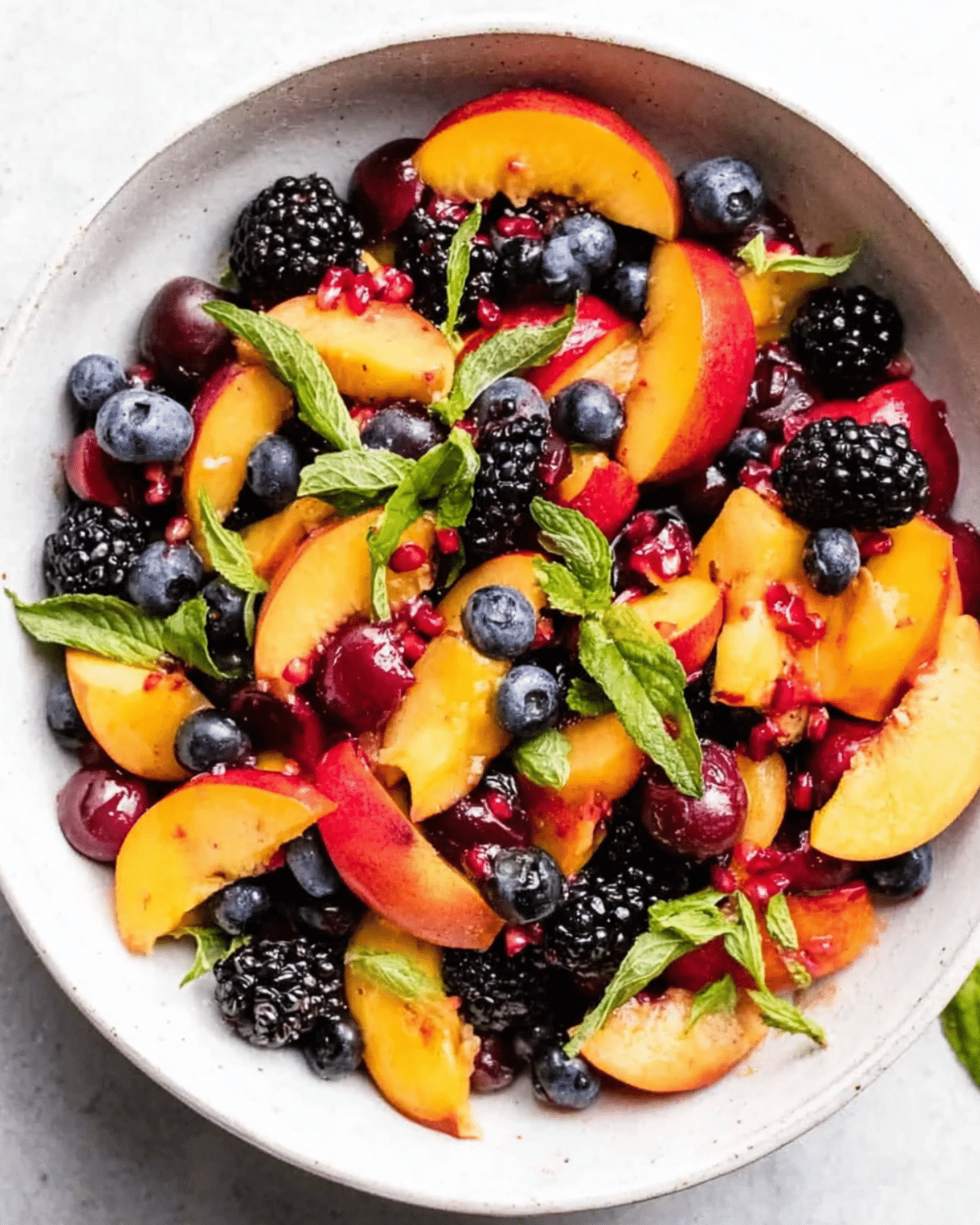A bowl of summer peach fruit salad with fresh peach slices, blueberries, blackberries, cherries, pomegranate seeds, and mint leaves.