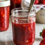 A clear glass jar filled with homemade strawberry freezer jam, with a spoon inside and fresh strawberries nearby.
