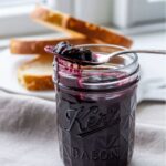 A jar of homemade blueberry butter with a spoonful of thick, glossy spread resting on the jar’s rim, slices of bread in the background.