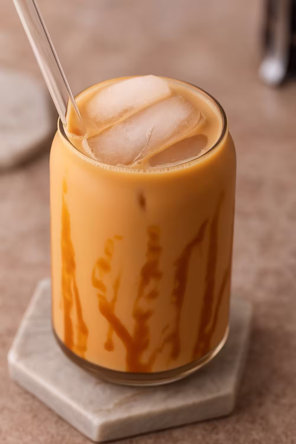 A glass of iced caramel coffee with ice cubes, creamy swirls, and caramel drizzle served with a clear glass straw.