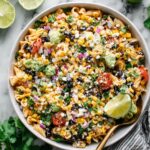 A vibrant bowl of Mexican Street Corn Pasta Salad with grilled corn, black beans, tomatoes, red onion, avocado, cotija cheese, and fresh cilantro, garnished with lime wedges.