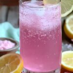 A refreshing glass of pink drink with ice, garnished with lemon slice, next to lemons and pink salt, representing a Pink Salt Weight Loss Recipe.