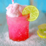 A vibrant pink Cotton Candy Margarita without alcohol, served in a mason jar with a slice of lime, pink cotton candy on top, and a pink-striped straw, surrounded by sugar crystals.
