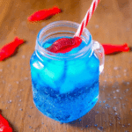 A mason jar filled with vibrant blue Sonic Ocean Water, topped with ice, a red Swedish Fish candy, and a red-and-white striped straw, on a wooden surface with water droplets and more candy fish around.