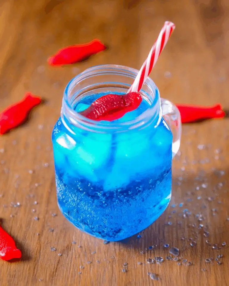 A mason jar filled with vibrant blue Sonic Ocean Water, topped with ice, a red Swedish Fish candy, and a red-and-white striped straw, on a wooden surface with water droplets and more candy fish around.