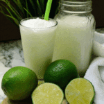 Brazilian lemonade with coconut served in a glass with ice, accompanied by fresh whole and halved limes, and a jar of the drink in the background.