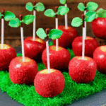 Apple Cake Pops with red sugar coating, candy leaves, and white sticks displayed on green grass-like base.