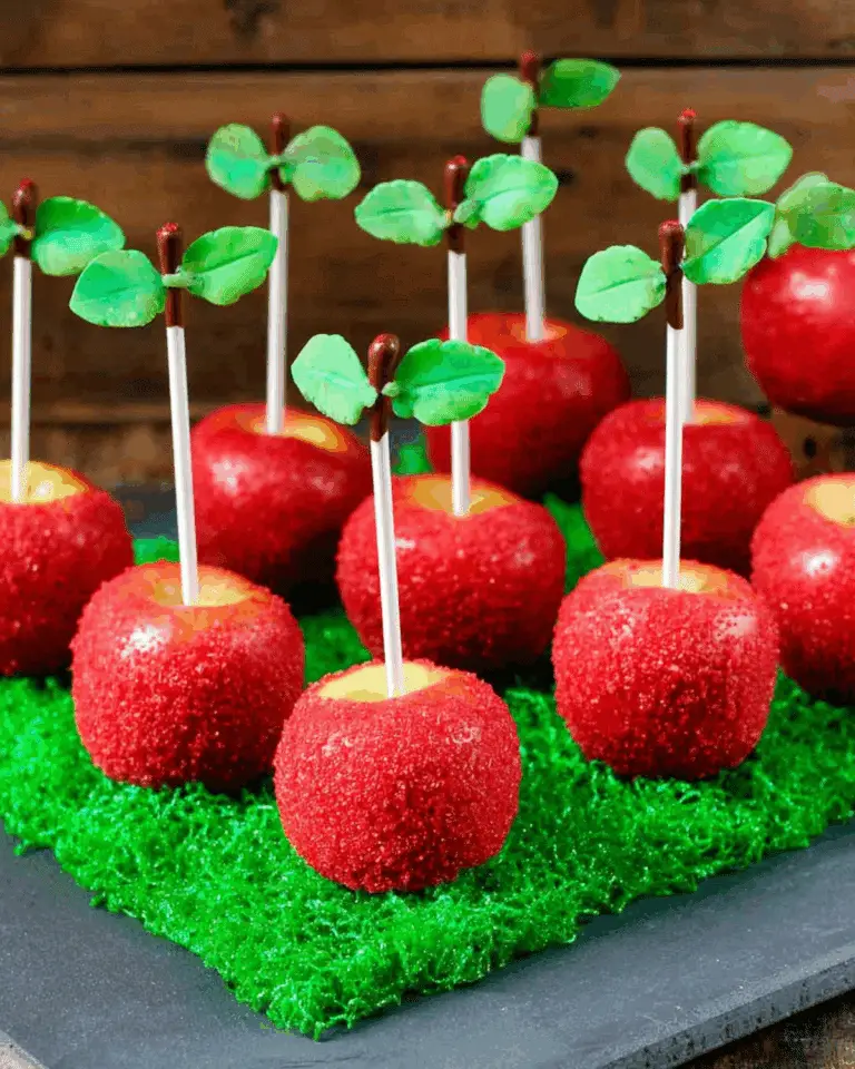 Apple Cake Pops with red sugar coating, candy leaves, and white sticks displayed on green grass-like base.