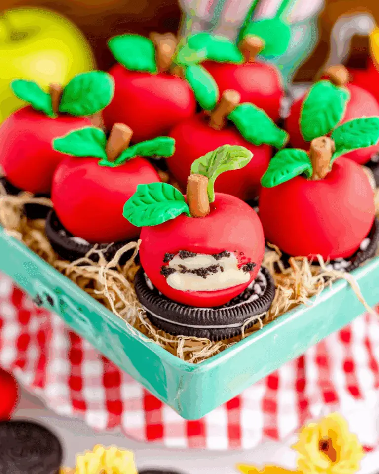 Decorative apple OREO balls covered in red coating with green fondant leaves and pretzel stems, placed on top of OREO cookies.