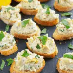 Artichoke bruschetta topped with creamy artichoke spread and fresh herbs on toasted baguette slices, served on a dark slate platter.