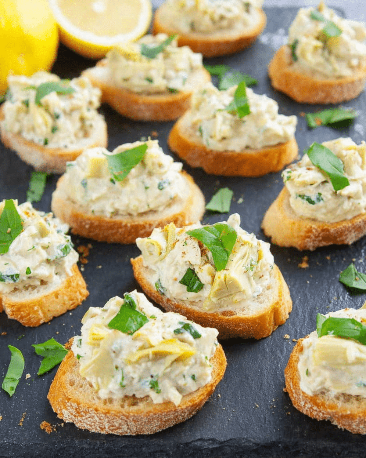 Artichoke bruschetta topped with creamy artichoke spread and fresh herbs on toasted baguette slices, served on a dark slate platter.