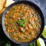 Bowl of fresh blender salsa garnished with parsley, surrounded by lime wedges, tortilla chips, and cilantro on a dark surface.