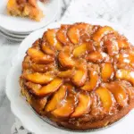 Bourbon peach upside down cake topped with caramelized peach slices, served on a white cake stand with a slice plated in the background.