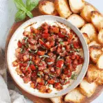 Bruschetta dip topped with diced tomatoes, basil, and balsamic glaze, served with toasted baguette slices on a wooden platter.