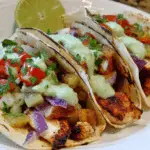 Three Chili Lime Chicken Tacos filled with grilled pineapple salsa, red onions, cilantro, and drizzled with creamy green sauce on a white plate with a lime wedge.