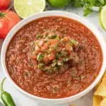 Bowl of Chili’s Copycat Salsa topped with diced tomatoes, onions, and cilantro, surrounded by fresh lime, tomatoes, and tortilla chips.