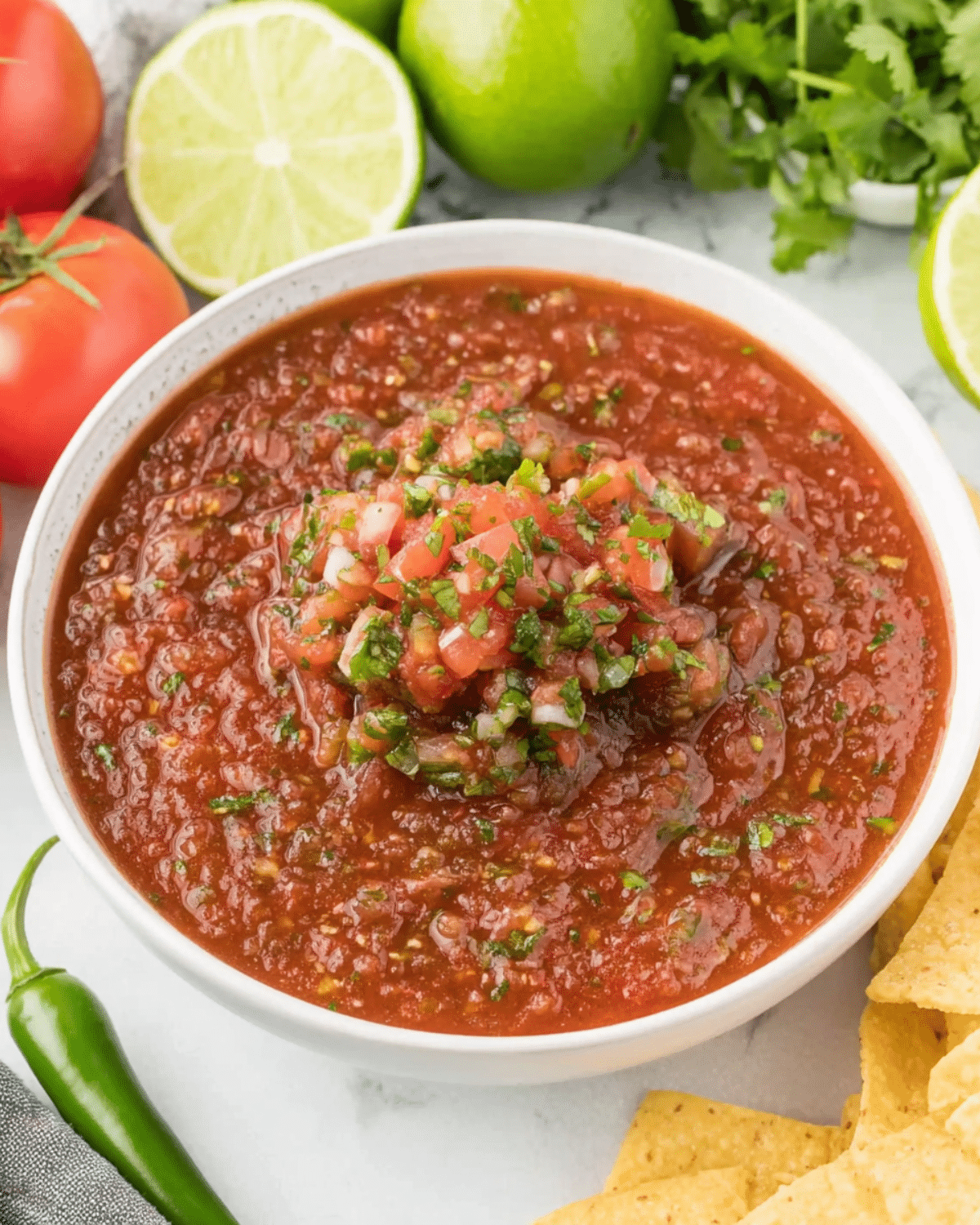 Bowl of Chili’s Copycat Salsa topped with diced tomatoes, onions, and cilantro, surrounded by fresh lime, tomatoes, and tortilla chips.
