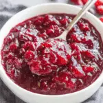 Classic homemade cranberry sauce in a white bowl with a spoon, showing a thick and chunky texture.