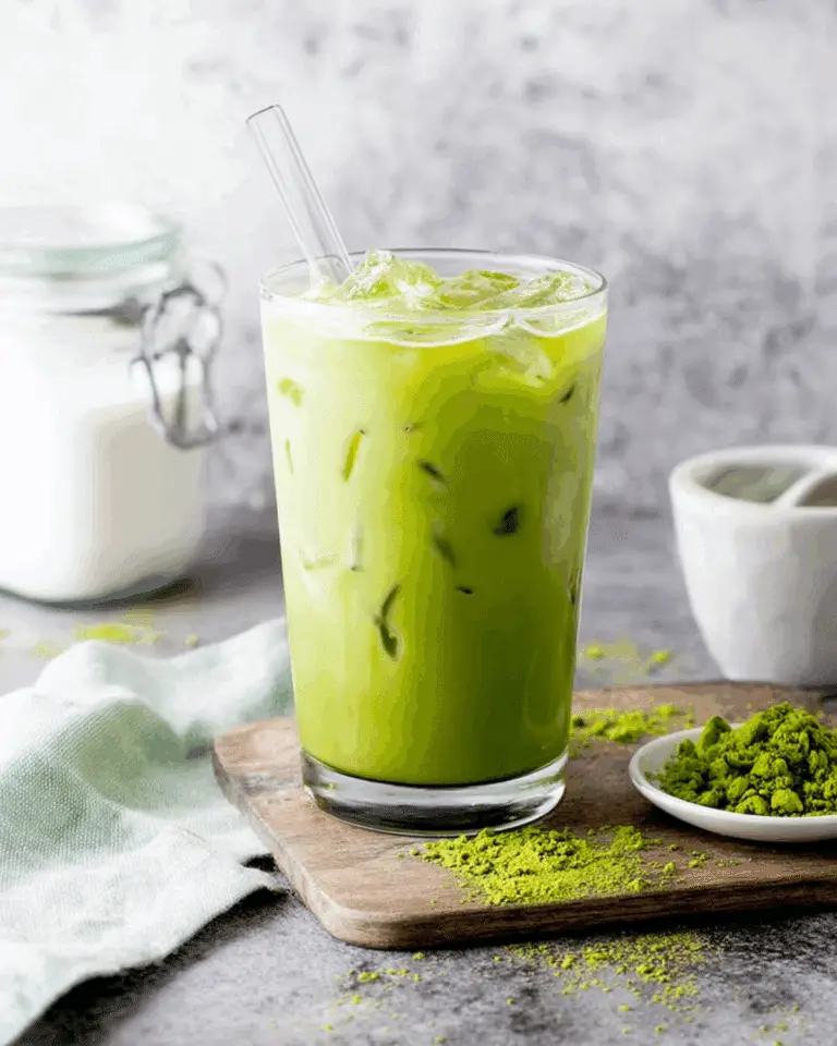 Glass of Copycat Starbucks Iced Matcha Latte with ice cubes and a clear straw, placed on a wooden board next to matcha powder and milk.