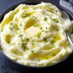 Creamiest baked mashed potatoes in a black bowl, topped with melted butter, chopped chives, and black pepper.