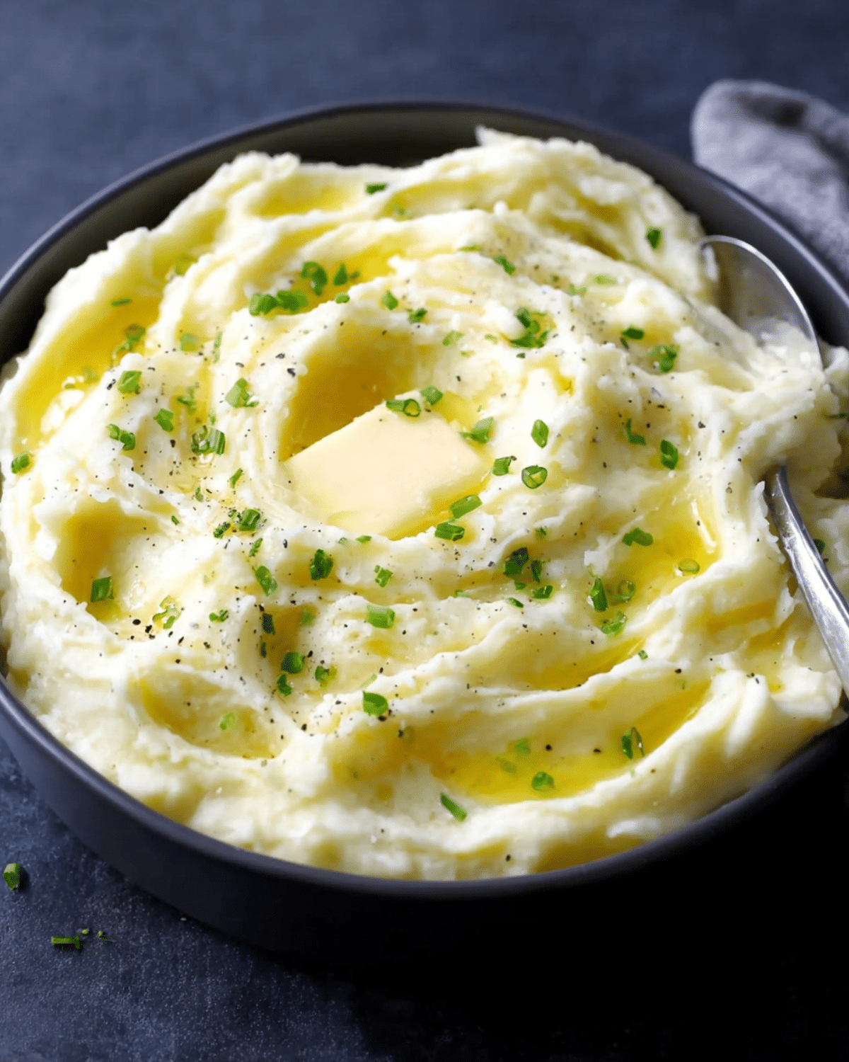 Creamiest baked mashed potatoes in a black bowl, topped with melted butter, chopped chives, and black pepper.