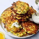 Crispy zucchini fritters stacked on a plate, served with a dollop of creamy herb sauce and a lemon wedge on the side.