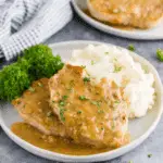 Plate of crock pot pork chops smothered in gravy, served with mashed potatoes and a side of parsley garnish.