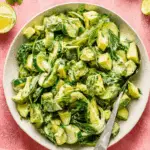 Bowl of cucumber avocado salad with creamy dressing, herbs, and lime on a pink background.