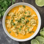 Bowl of curry Thai dumpling soup with plump dumplings in a creamy, orange curry broth, garnished with chopped cilantro and lime slices on the side.