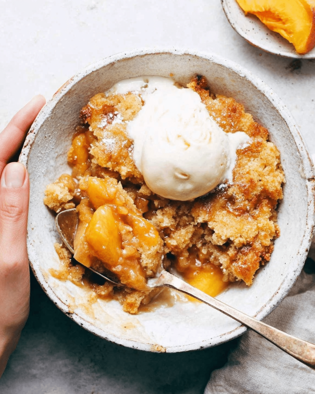 Bowl of fresh peach cobbler topped with a scoop of vanilla ice cream, featuring golden baked crust and juicy peach filling.