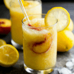 Frozen Peach Lemonade Slushie in a clear glass, garnished with a lemon slice and fresh peach inside.