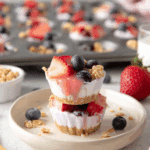 Frozen Yogurt Bites topped with strawberries, blueberries, and granola, stacked on a plate with more bites in a muffin tray in the background.