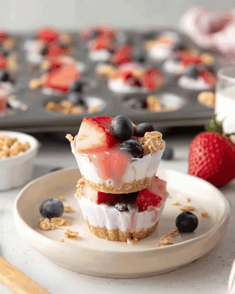 Frozen Yogurt Bites topped with strawberries, blueberries, and granola, stacked on a plate with more bites in a muffin tray in the background.