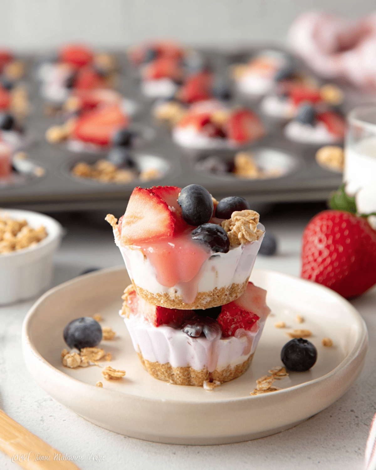 Frozen Yogurt Bites topped with strawberries, blueberries, and granola, stacked on a plate with more bites in a muffin tray in the background.