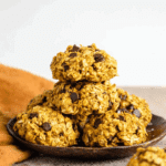 Healthy pumpkin oat cookies with chocolate chips stacked on a dark plate, with an orange napkin in the background.