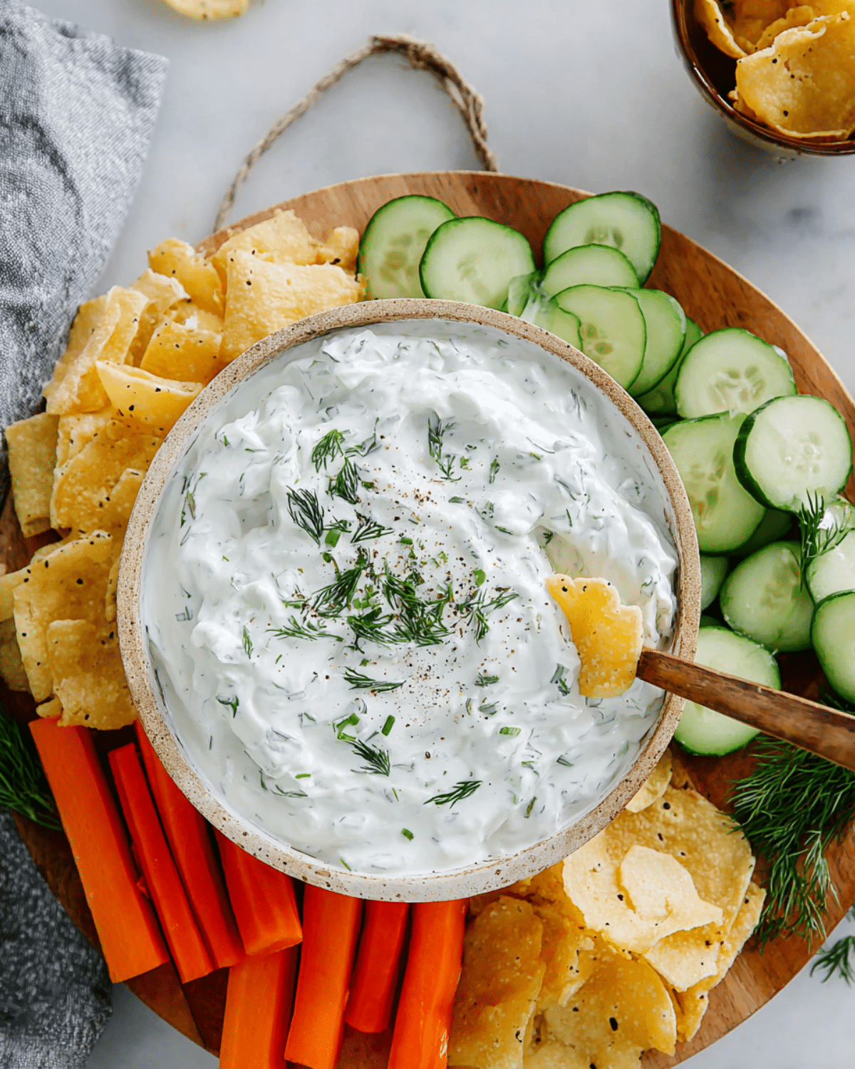 Bowl of creamy Homemade Pickle Dip garnished with fresh dill, surrounded by cucumber slices, carrot sticks, and chips on a wooden serving board.