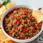 Bowl of homemade salsa recipe with fresh tomatoes, surrounded by tortilla chips, with a hand dipping a chip into the salsa.