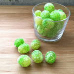 Kool-Aid grapes coated in sugar, displayed in a clear glass with several scattered on a wooden surface.