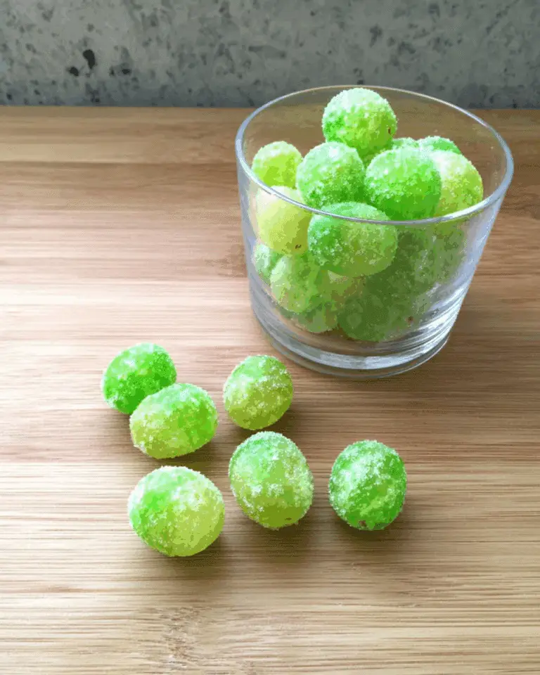 Kool-Aid grapes coated in sugar, displayed in a clear glass with several scattered on a wooden surface.