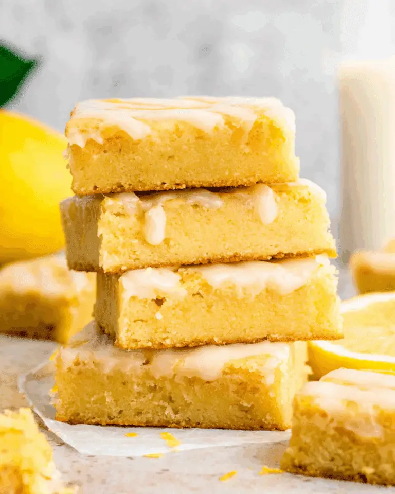 Lemon Brownie Bars stacked on parchment paper, drizzled with lemon glaze and surrounded by fresh lemon slices.
