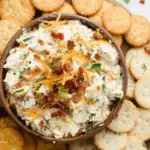 Bowl of creamy Million Dollar Dip topped with shredded cheddar cheese and bacon bits, surrounded by buttery round crackers.