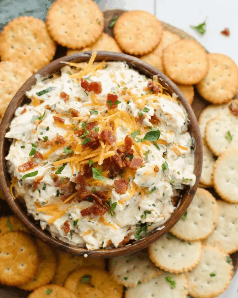 Bowl of creamy Million Dollar Dip topped with shredded cheddar cheese and bacon bits, surrounded by buttery round crackers.