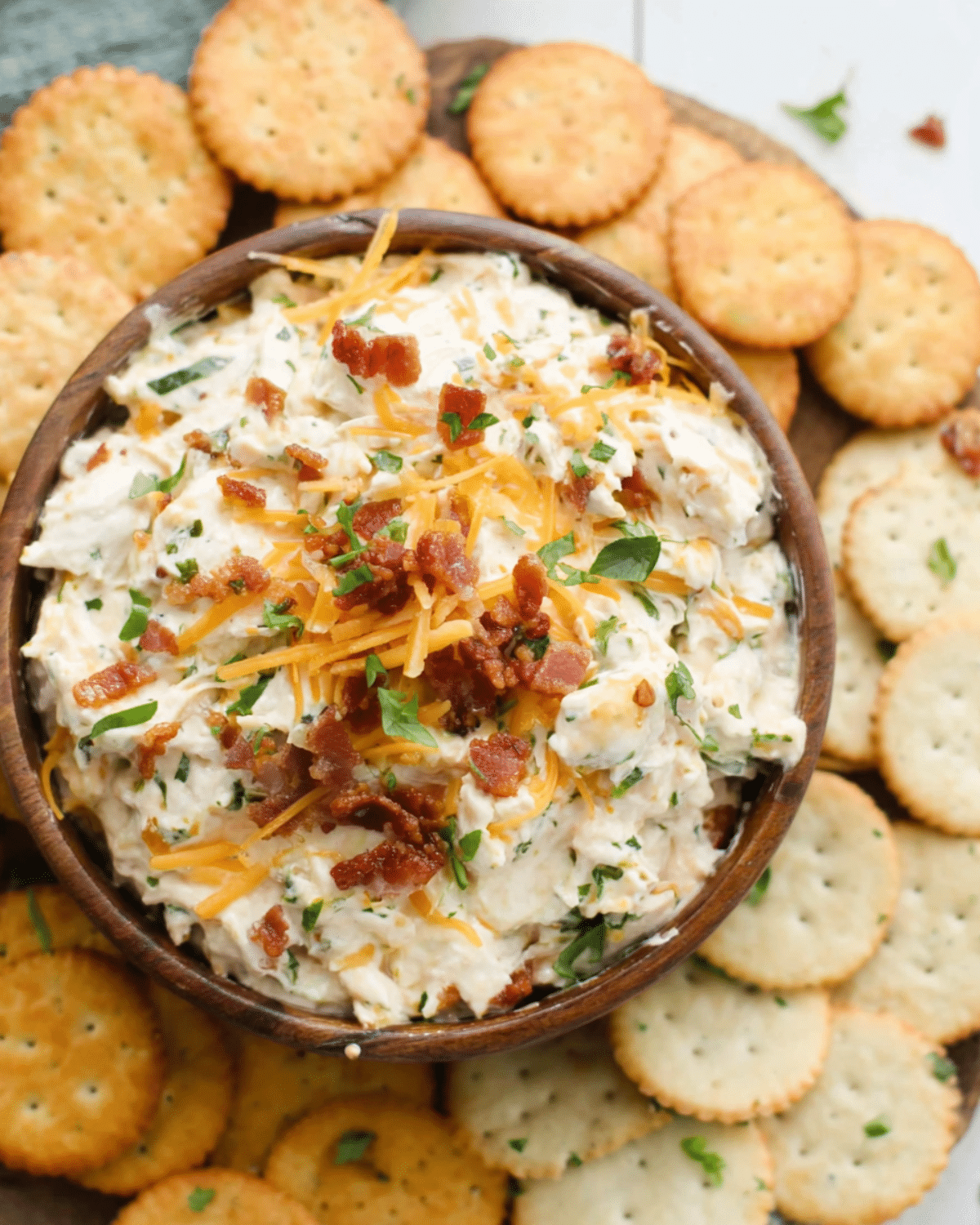 Bowl of creamy Million Dollar Dip topped with shredded cheddar cheese and bacon bits, surrounded by buttery round crackers.